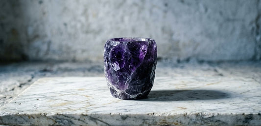 A hand-carved solid amethyst drinking vessel with deep imperial violet tones and natural white veining, centered on an aged white marble slab.